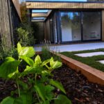 Lush green garden foliage surrounding a Vivid Green garden room with anthracite grey sliding doors and windows, open overhang and composite decked seating area, and stepping stones leading to the entrance
