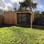 Vivid Green garden room with western red cedar cladding, anthracite grey sliding door and window and an adjacent compsite decking area featuring furniture, situated at the bottom of a grassy garden with a small tree in front of the room.