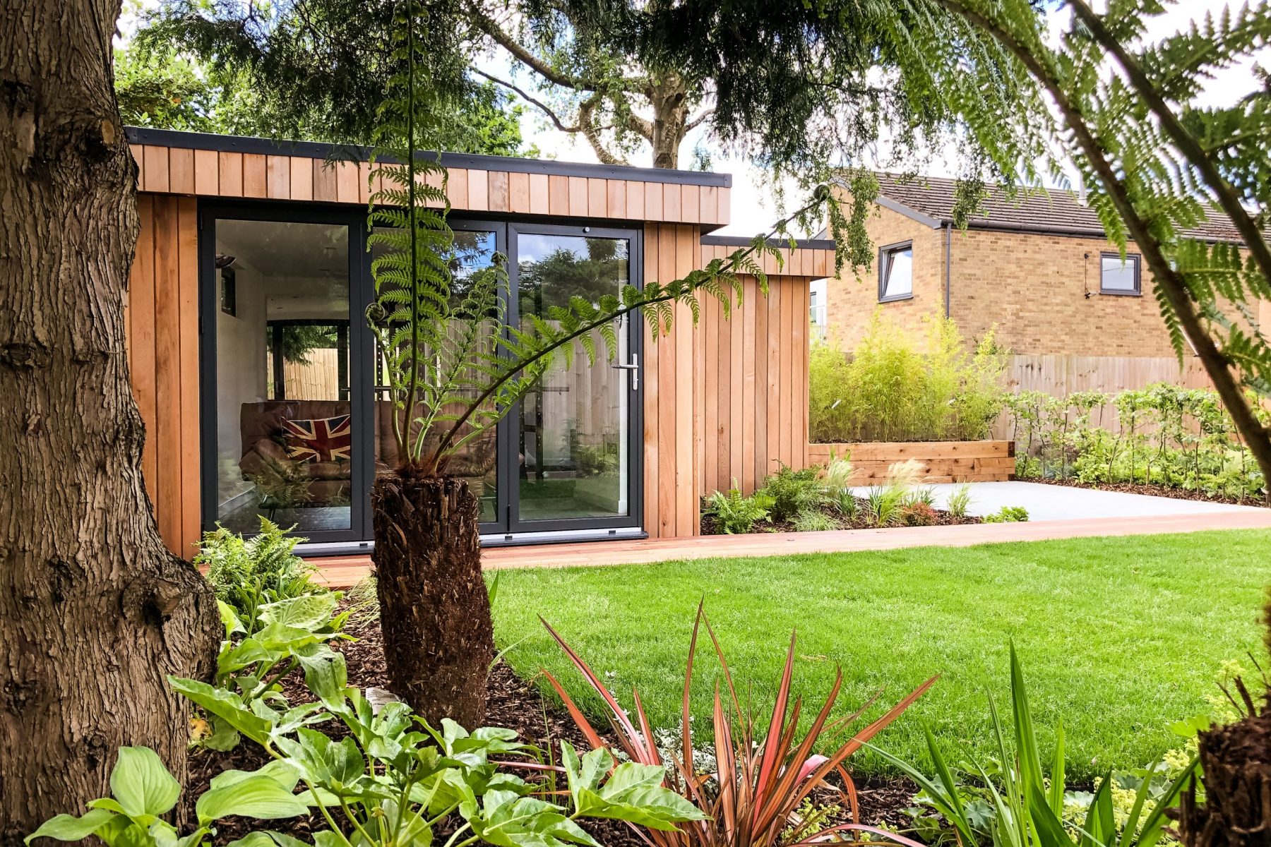 Vivid Green western red cedar clad, L shaped garden office with anthracite grey aluminium bi-fold doors, a concrete and bricked patio to the right of it and lush foliage in foreground.