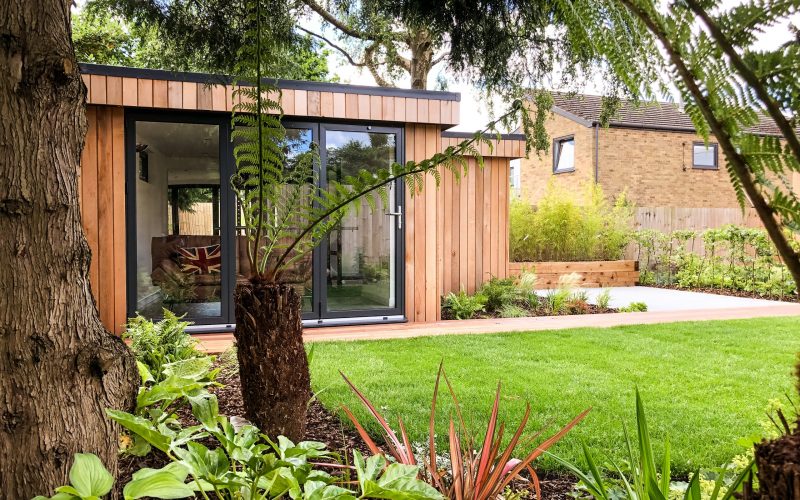 Vivid Green western red cedar clad, L shaped garden office with anthracite grey aluminium bi-fold doors, a concrete and bricked patio to the right of it and lush foliage in foreground.