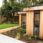 Anthracite grey framed glass windowed, western red cedar clad Vivid Green l-shaped garden studio, around a tree with a decked walkway and furniture on a rainy day