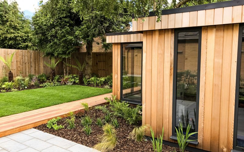 Anthracite grey framed glass windowed, western red cedar clad Vivid Green l-shaped garden studio, around a tree with a decked walkway and furniture on a rainy day