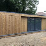 Vivid Green garden room with thermowood cladding, grey French doors and full height fixed screens, featuring a closed storage room with secret door and composite deck step, situated on a natural dirt surface with trees visible in the background.
