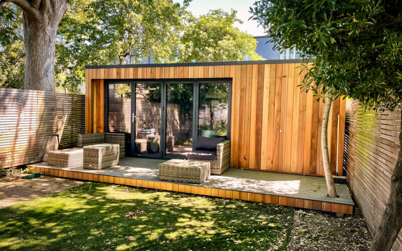 Exterior view of a flat-roofed Vivid Green garden room with western red cedar cladding, anthracite grey bifold doors and windows, featuring an elevated composite grey deck with patio furniture in front of the entrance, surrounded by a garden and wooden neighboring walls.
