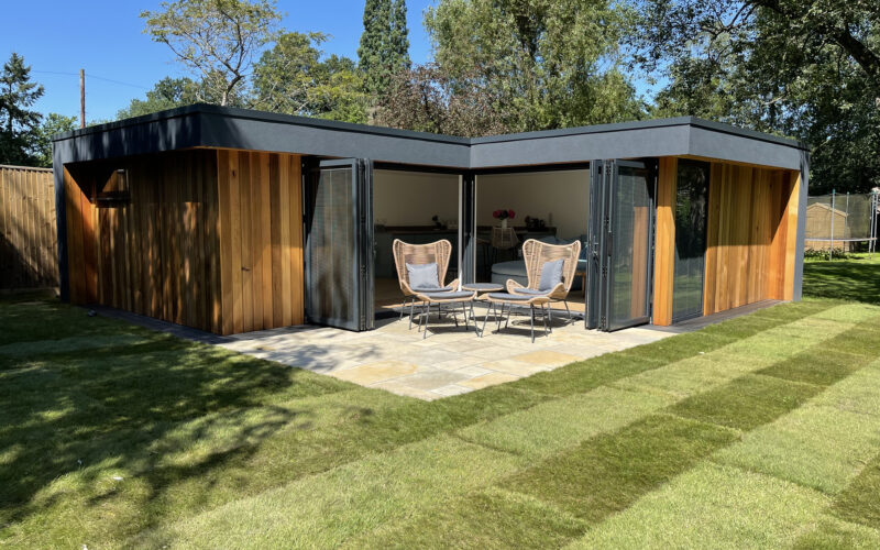 Exterior view of a Vivid Green L-shaped garden studio with a flat roof, western red cedar cladding, grey rendered overhang, anthracite grey aluminium bi-fold doors opening onto a small tiled patio, and furniture set on it, surrounded by an artificial grass garden on a sunny day.
