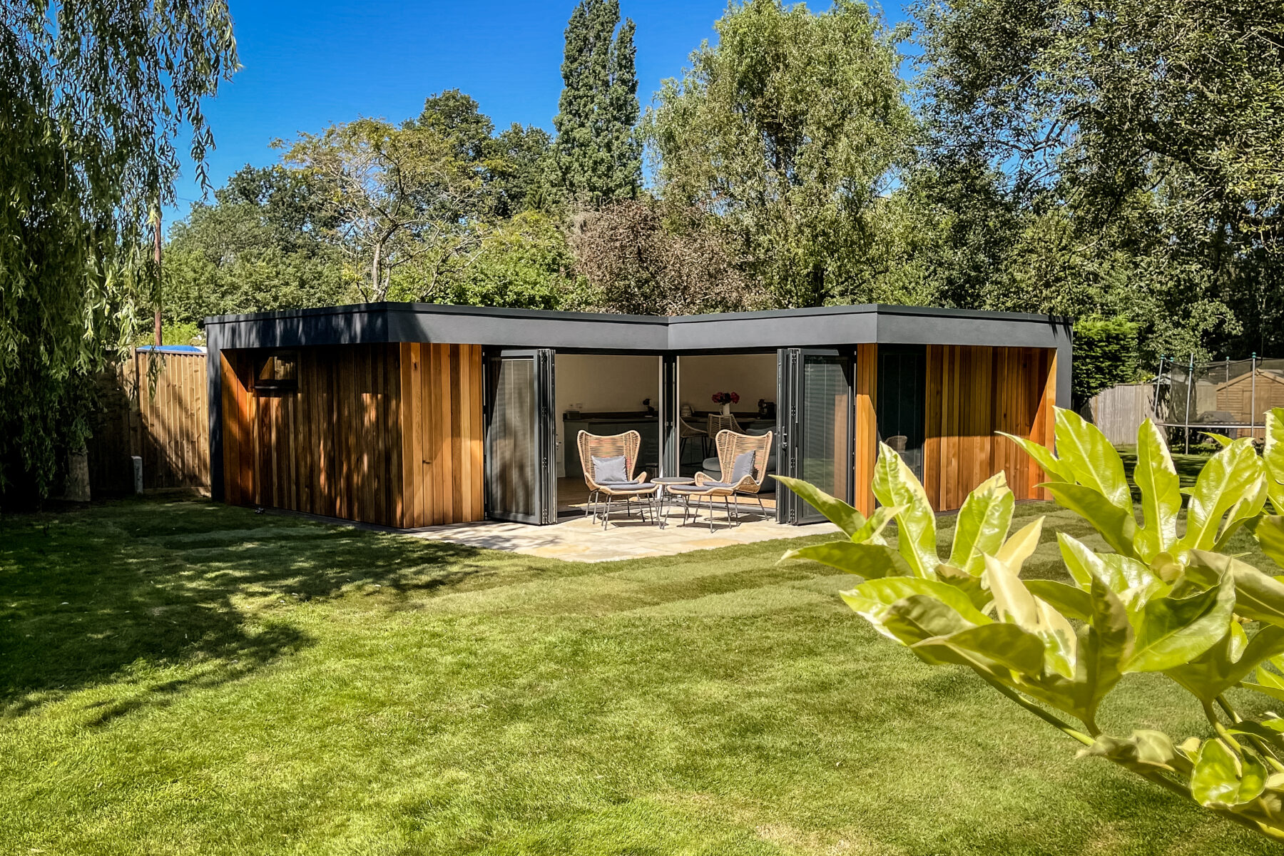 Exterior view of a Vivid Green L-shaped garden room with a flat roof, anthracite grey aluminium bi-fold doors opening onto a small tiled patio, and furniture set on it, surrounded by foliage and an artificial grass garden on a sunny day