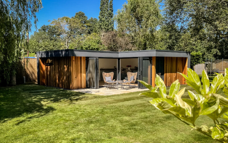 Exterior view of a Vivid Green L-shaped garden room with a flat roof, anthracite grey aluminium bi-fold doors opening onto a small tiled patio, and furniture set on it, surrounded by foliage and an artificial grass garden on a sunny day
