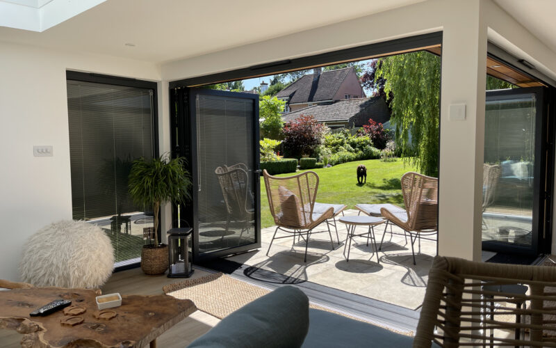 Interior of a furnished L-shaped garden room with anthracite grey aluminium bi-fold doors with integral blinds opening onto a tiled patio with outdoor furniture and a sunny grass lawn, with the main house visible in the background.