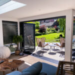Interior of a furnished L-shaped garden room with anthracite grey bi-fold doors with integral blinds opening onto a tiled patio with outdoor furniture and a sunny grass lawn, with the main house visible in the background.