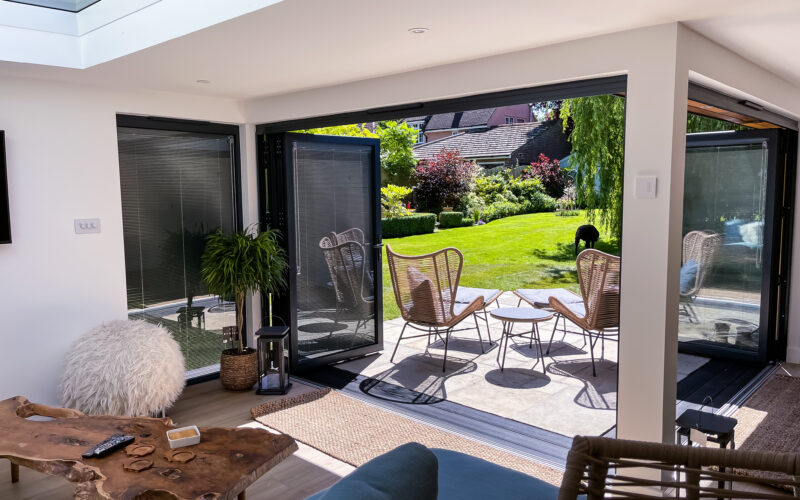 Interior of a furnished L-shaped garden room with anthracite grey bi-fold doors with integral blinds opening onto a tiled patio with outdoor furniture and a sunny grass lawn, with the main house visible in the background.