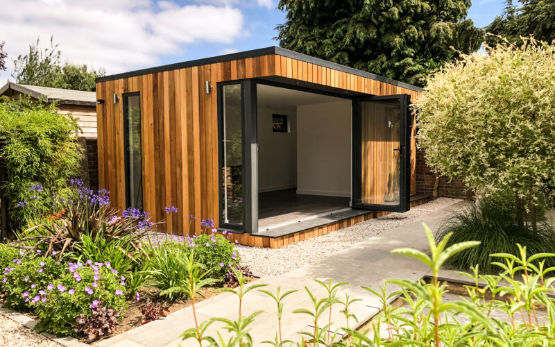Exterior view of an unfurnished Vivid Green flat-roofed garden room with open grey bi-fold doors in a gravelled backyard surrounded by foliage and a concrete walkway leading up to the entrance.
