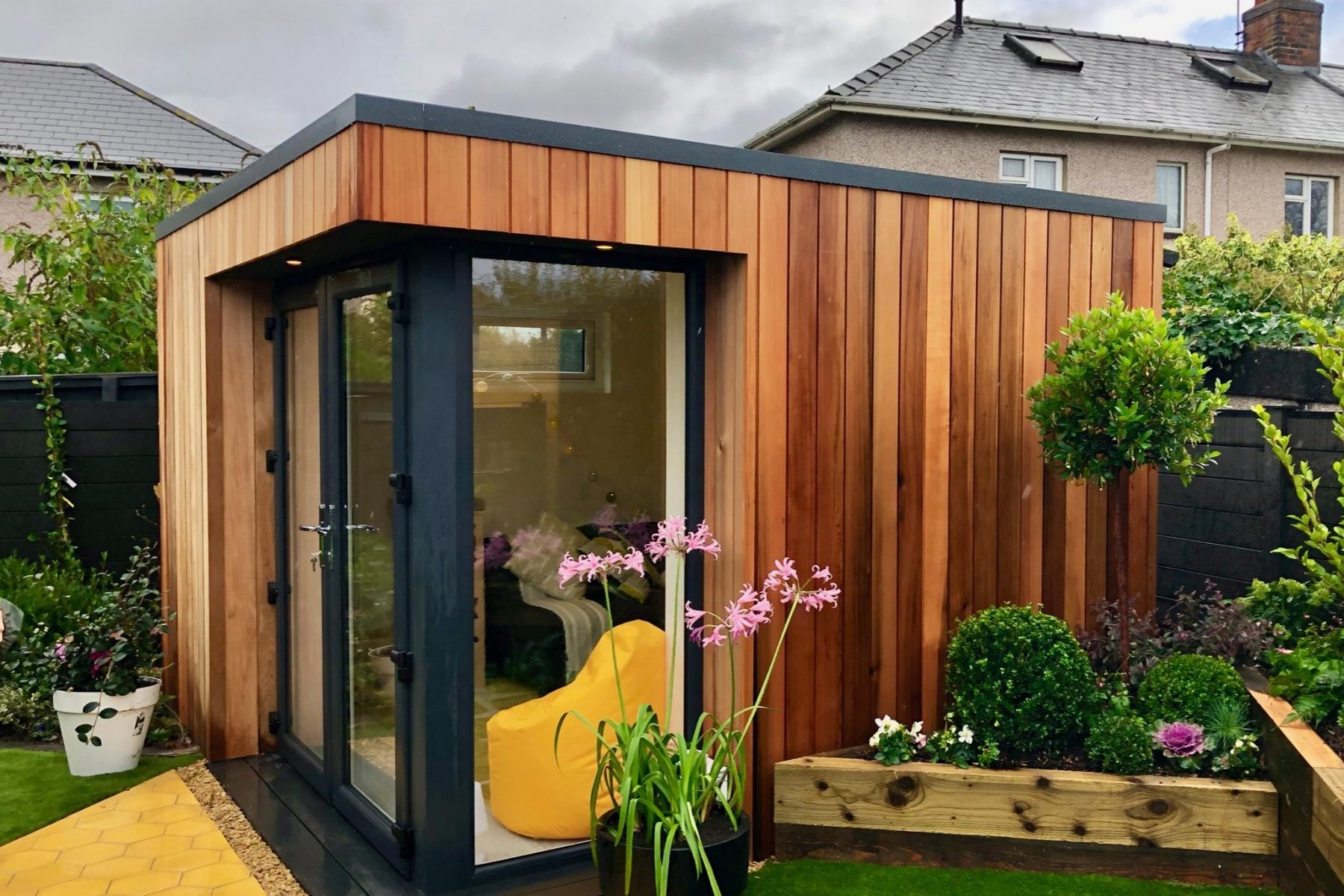 Furnished Vivid Green garden room with western red cedar cladding, anthracite grey corner glass door and windows, yellow beanbag, and surrounded by pot plants, foliage and a yellow walkway.