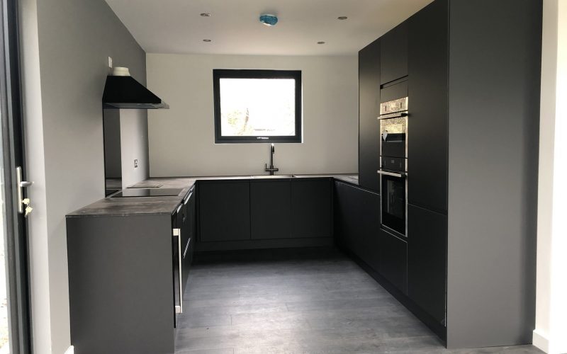 Black kitchen in Vivid Green SIPs annexe featuring built in cupboards, extractor fan, window over sink double oven and charcoal wood flooring