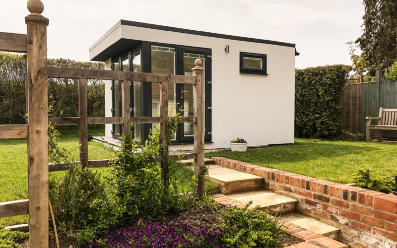 Vivid Green garden office with white render cladding and a flat roof, featuring anthracite grey doors and windows. Brick steps lead up to the entrance, while a growing garden is visible in the foreground and a garden bench in the background