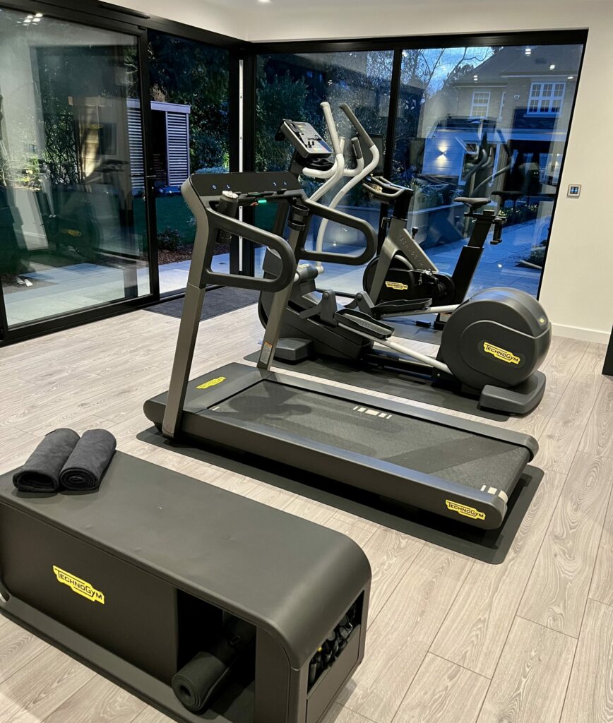 Interior view of Vivid Green garden gym studio with anthracite grey aluminium framed corner glass sliding doors looking out into the dark, ceiling down lights and four pieces of black gym equipment