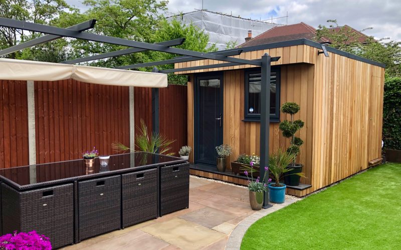Small Vivid Green garden office space with western red cedar anthracite grey door and window. Potted plants outside the door, and a tiled patio with an outdoor dining set and overhead pergola and a grass garden off to the side.