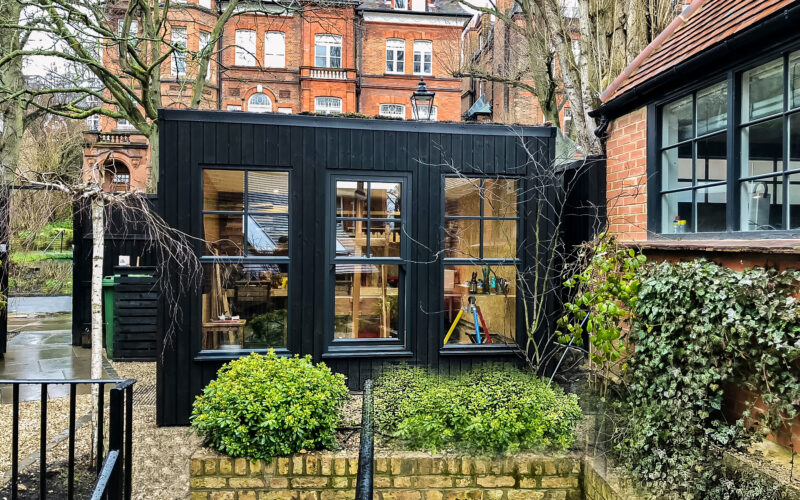 Exterior view of the Vivid Green art studio with stained black larch cladding, black framed timber sash windows, and a flat roof in a gravelled courtyard surrounded by green plants, trees, and neighboring buildings to the right and in the background.