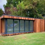 Vivid Green garden room with closed storage room, clad in oiled western red cedar, anthracite grey aluminium bi-fold doors, flat roof, and a view of trees in the background and grassy garden in the foreground.