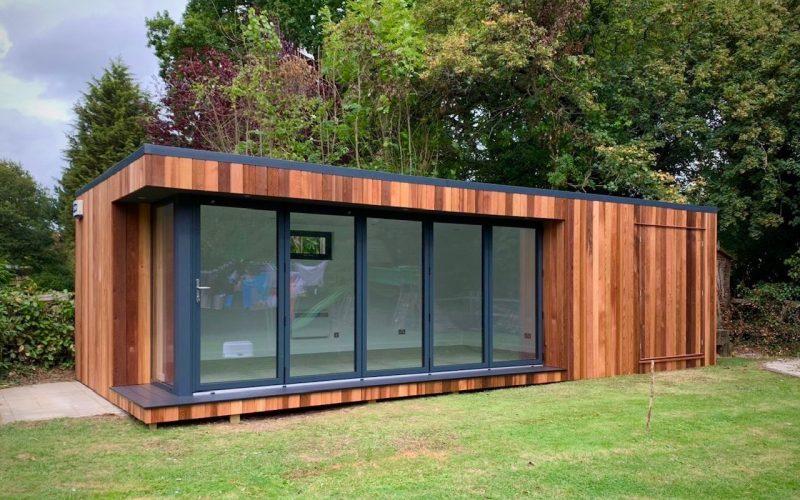 Vivid Green garden room with closed storage room, clad in oiled western red cedar, anthracite grey aluminium bi-fold doors, flat roof, and a view of trees in the background and grassy garden in the foreground.