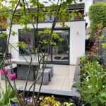 Vivid Green white rendered SIPs House Extension with outdoor dining set on tiled deck, surrounded by lush foliage, with garden room in background featuring glass sliding doors