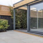 Exterior view of large garden office with anthracite grey sliding doors and tiled patio, situated close to a black and tan wall with a creeper plant growing on it.