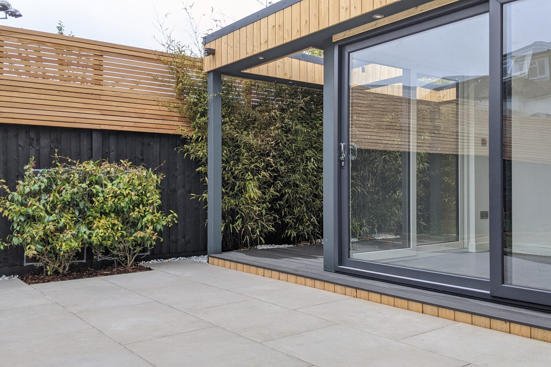 Exterior view of large garden office with anthracite grey sliding doors and tiled patio, situated close to a black and tan wall with a creeper plant growing on it.