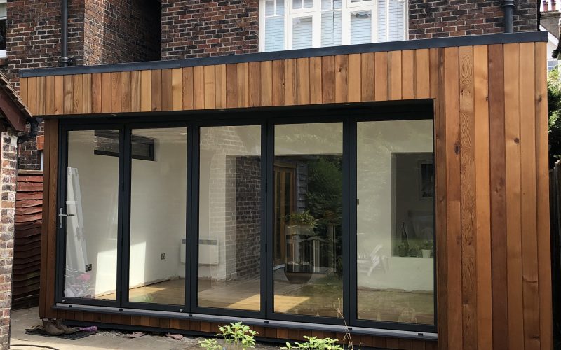 Exterior view of a Vivid Green flat-roofed SIPs house extension at the rear of a house, with anthracite grey bi-fold doors giving a glimpse of the nearly complete interior.