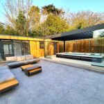 Vivid Green garden office space with flat roof, western red cedar cladding, anthracite grey aluminium doors and windows, featuring modern outdoor furniture set and a large pergola with frameless sliding doors covering a big Jucuzzi