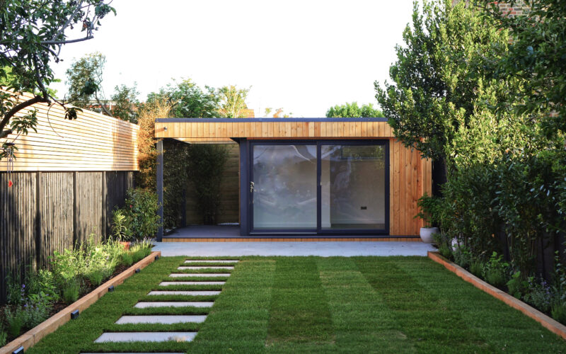 Vivid Green garden office at bottom of grass garden, with western red cedar cladding, anthracite grey sliding corner doors, large decking and overhang. Neighbouring wall to the left, and green hedge to right with stepping stones leading up to the entrance.