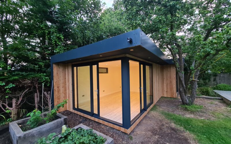 Small unfurnished Vivid Green L-shaped garden office space with open anthracite grey corner sliding doors nestled at the bottom of a garden next to pot plants and a tree with the lights on illuminating the interior.