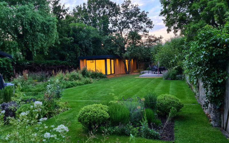 Exterior view of a Vivid Green garden office space situated at the bottom of a lush garden with lights illuminating the interior during late afternoon