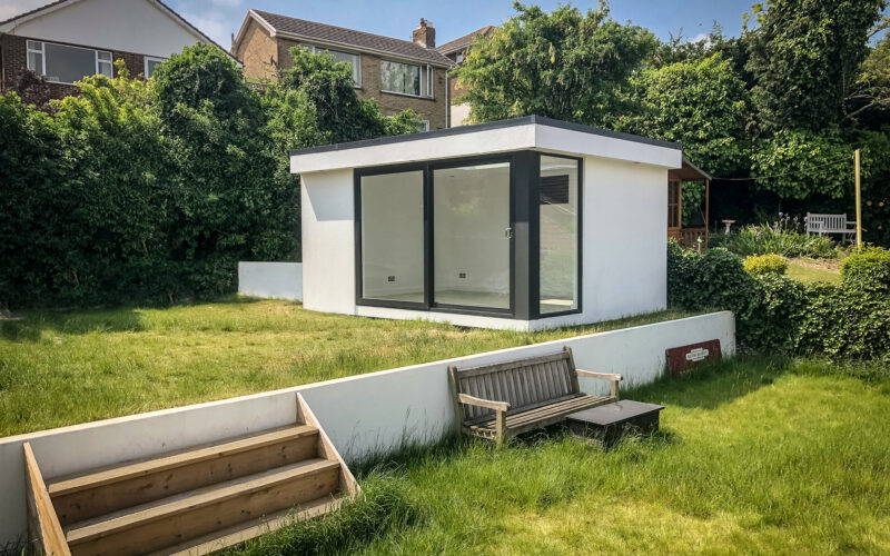 Small Vivid Green garden office space with white rednered exterior cladding, flat roof and anthracite grey sliding doors and corner window situated on the second level of the garden above a small white wall and garden bench.