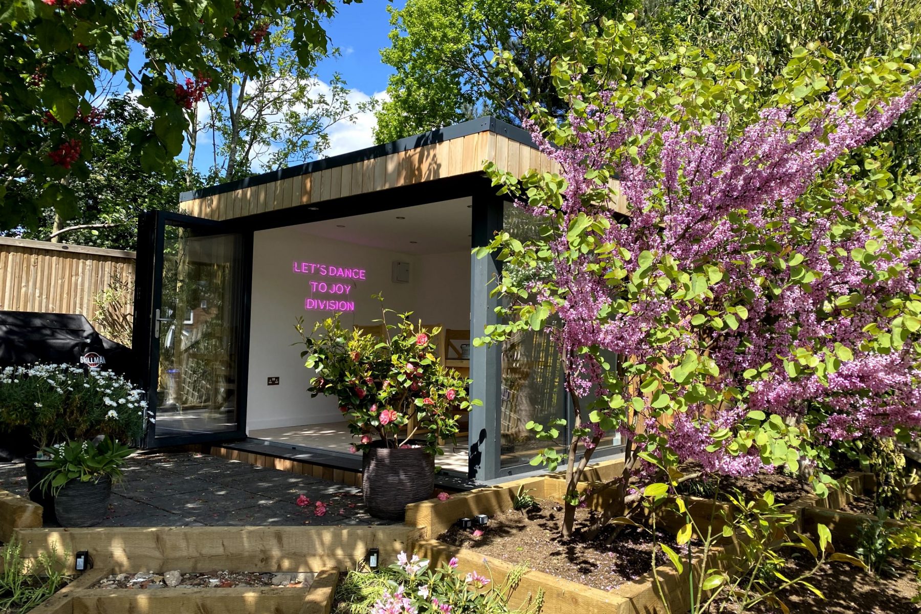 Vivid Green garden room with anthracite grey aluminium bi-fold doors meeting on the corner, clad in larch with large decking area to enjoy the lush garden in which it is set.
