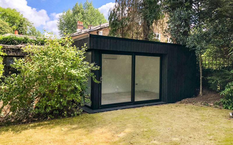 Exterior view of a black clad Vivid Green small garden office space featuring black framed sliding doors, situated at the bottom of a lush garden