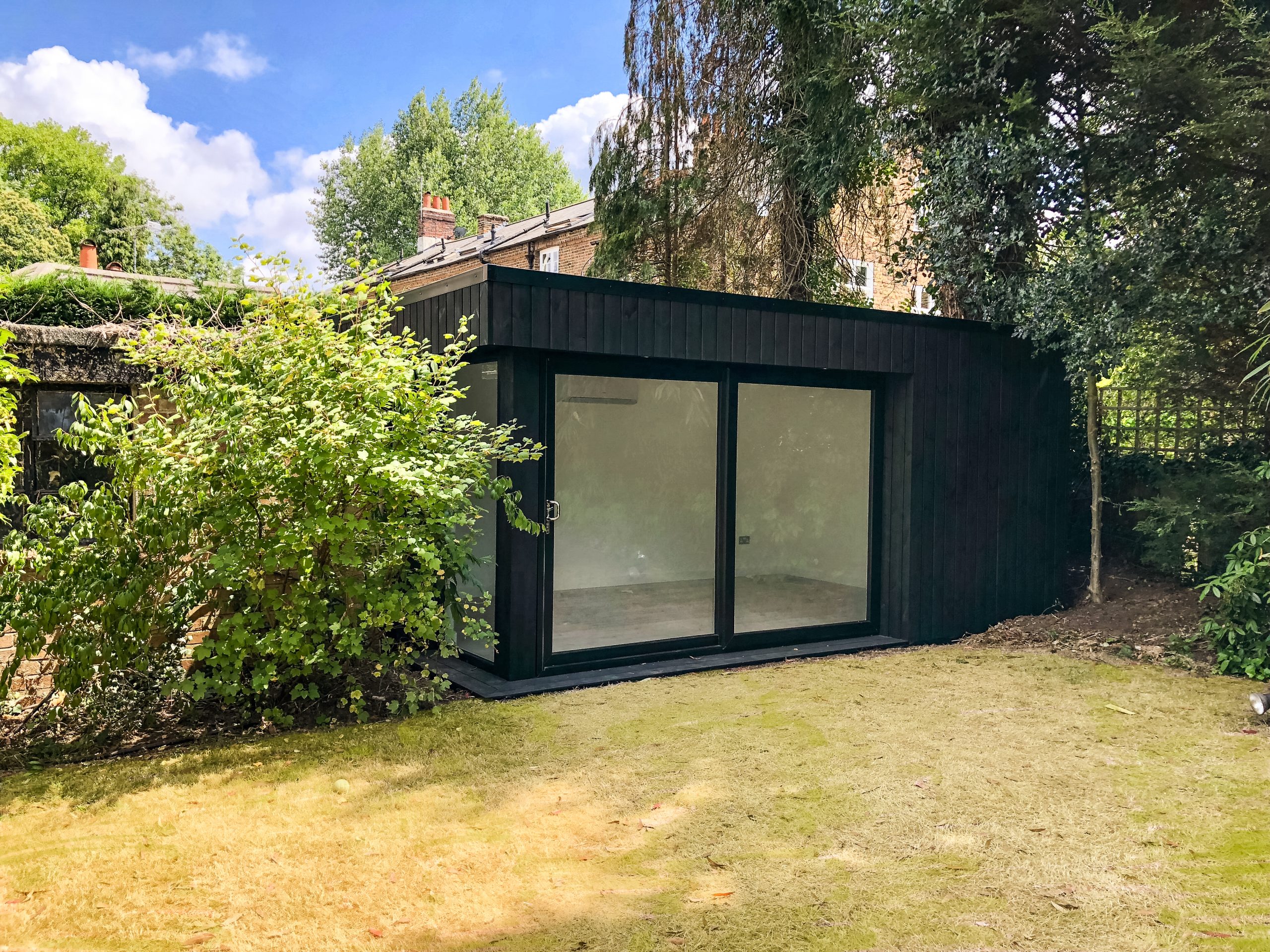 Exterior view of a black clad Vivid Green small garden office space featuring black framed sliding doors, situated at the bottom of a lush garden