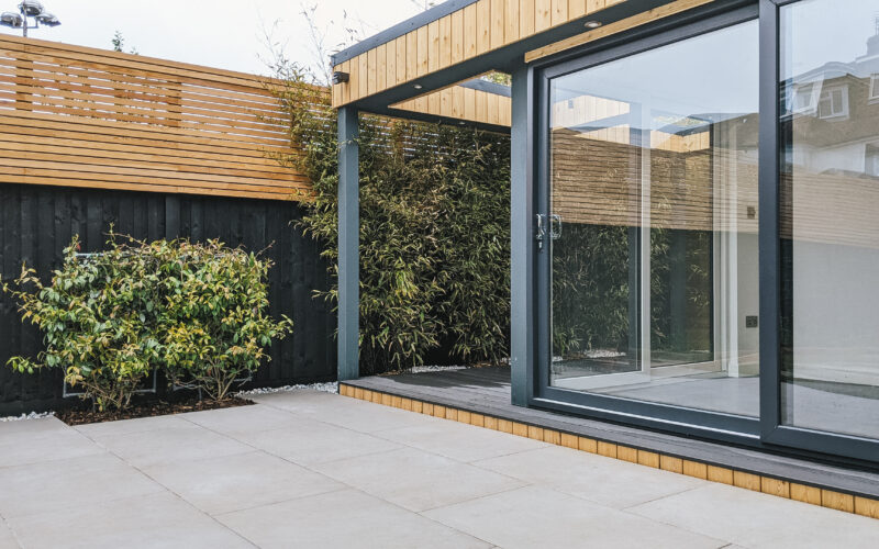 Exterior view of a large Vivid Green garden office with anthracite grey aluminium sliding doors and tiled patio, situated close to a black and tan wall with a creeper plant growing on it.