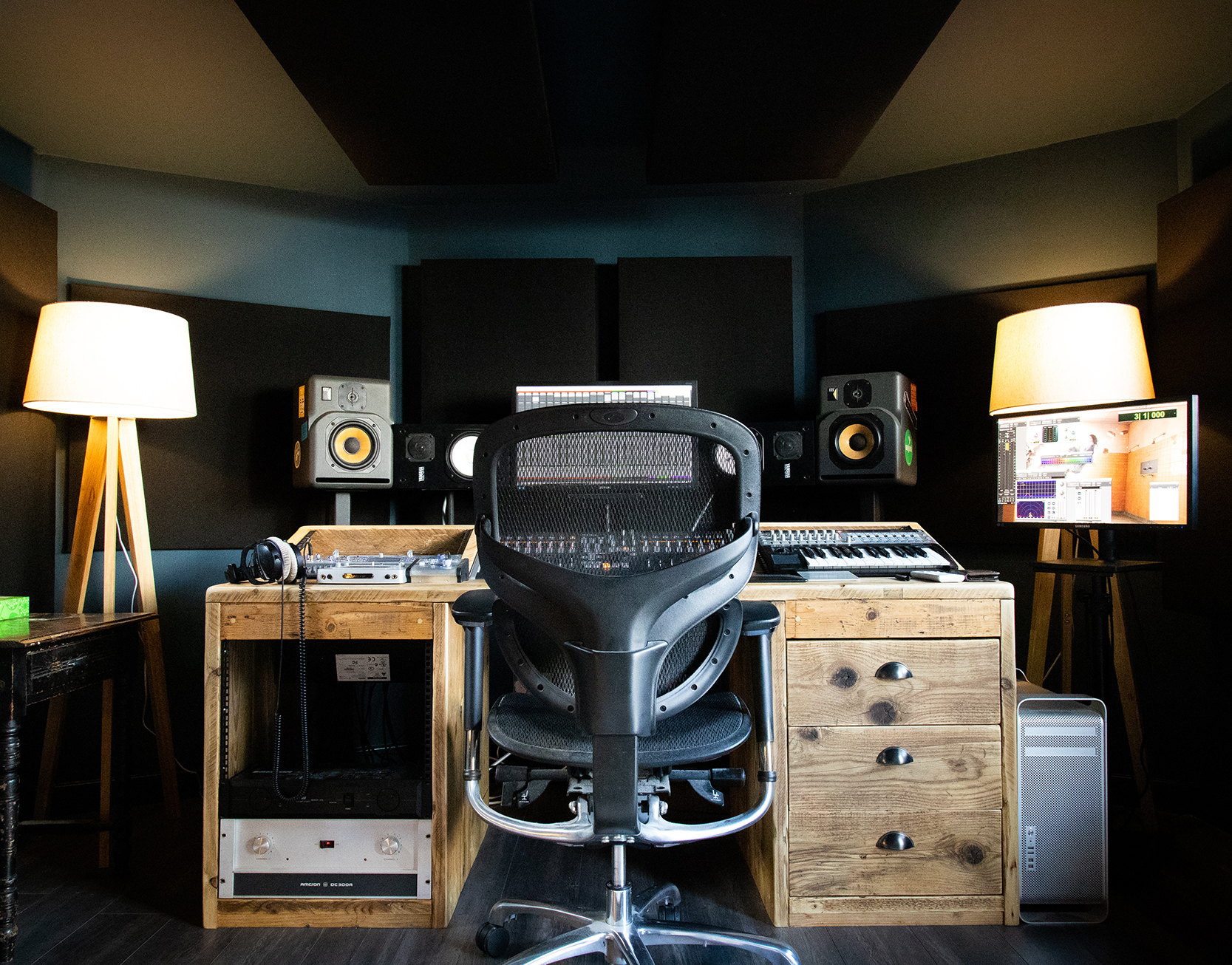Full view of a Vivid Green garden music studio's desk and technical equipment with a screen and speakers mounted above, warm light lamps on either side, and soundproofing on walls and roof.