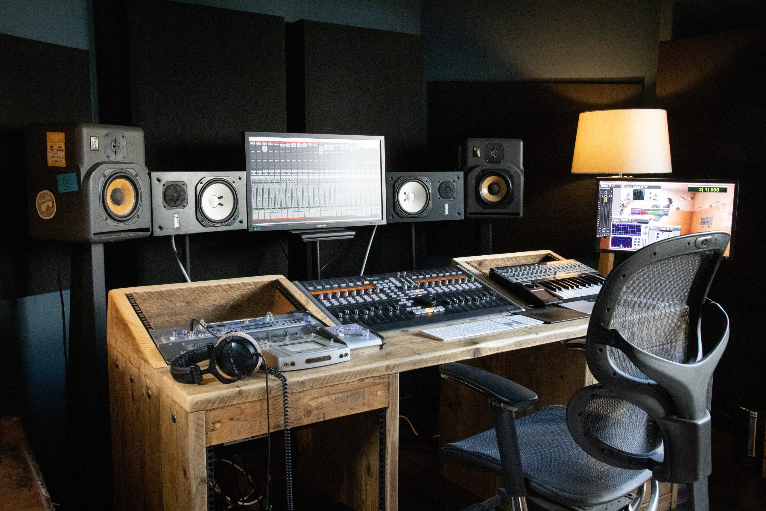 Interior view of a Vivid Green home music studio's desk with technical equipment, an empty black office chair, speakers, and a screen mounted above the desk, accompanied by a warm light lamp on the right side.