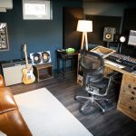 Vivid Green music studio with production equipment, including a screen music program, speakers, mixing desk, keyboard, and a standing lamp, furnished with a tan couch with white cushions and carpet against the opposite wall
