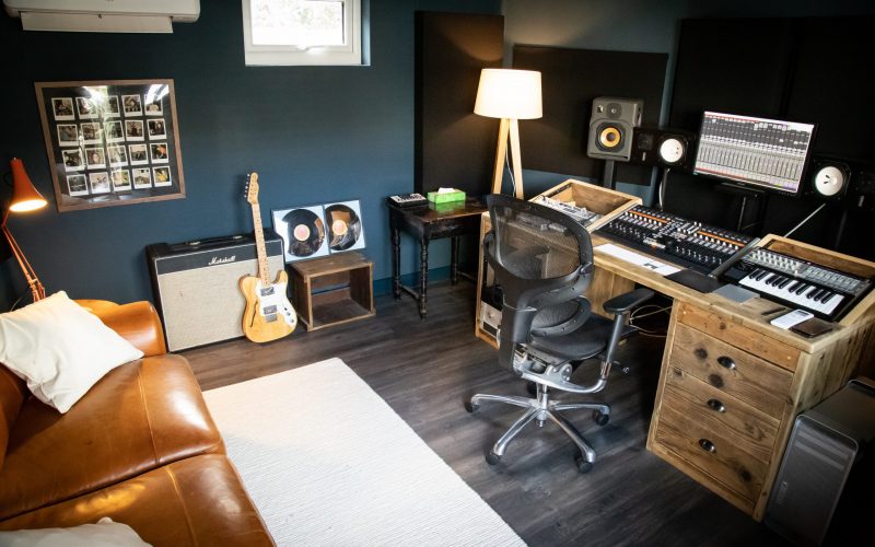 Vivid Green music studio with production equipment, including a screen music program, speakers, mixing desk, keyboard, and a standing lamp, furnished with a tan couch with white cushions and carpet against the opposite wall
