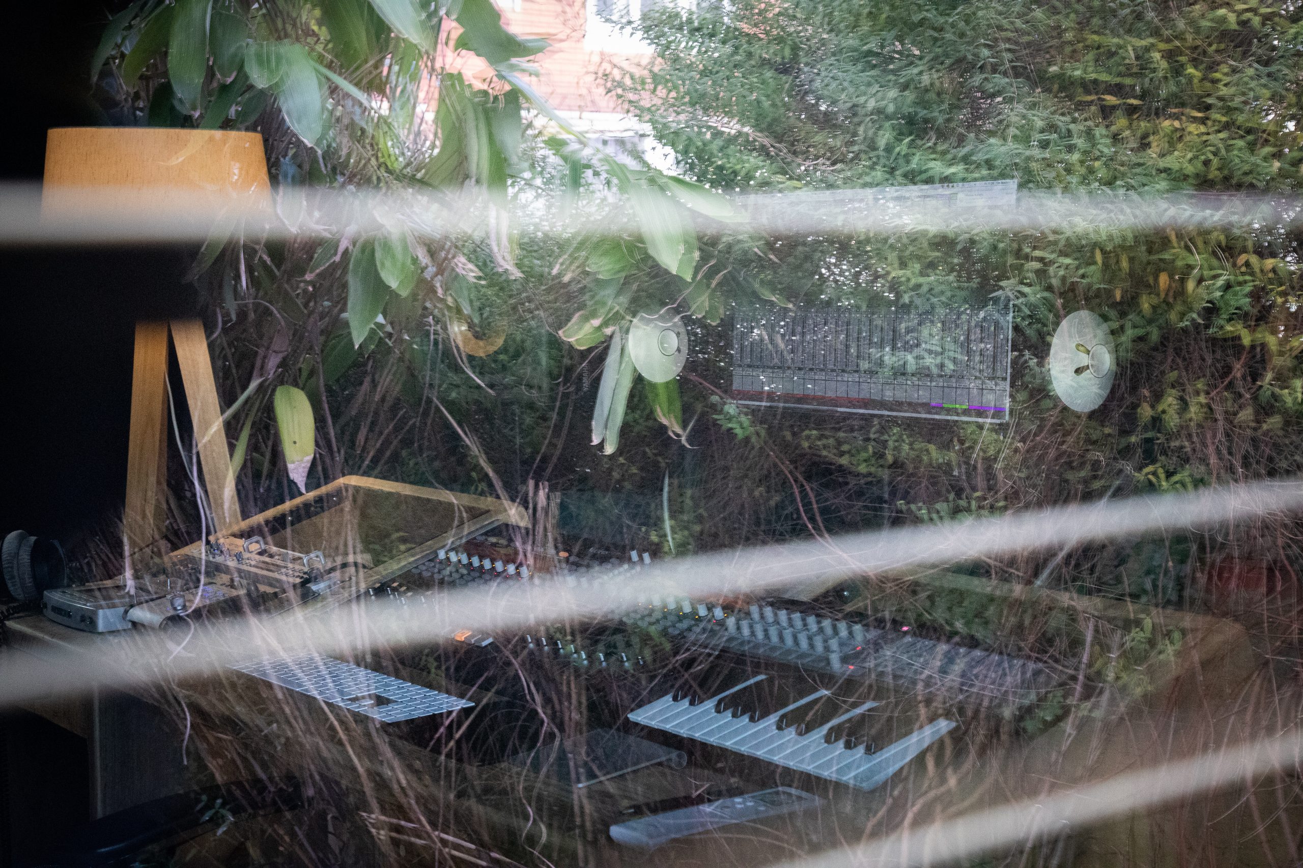 Blurred view of the backyard as seen through the open blinds of a Vivid Green home music studio window, with the reflection of the music equipment from inside visible on the glass pane.