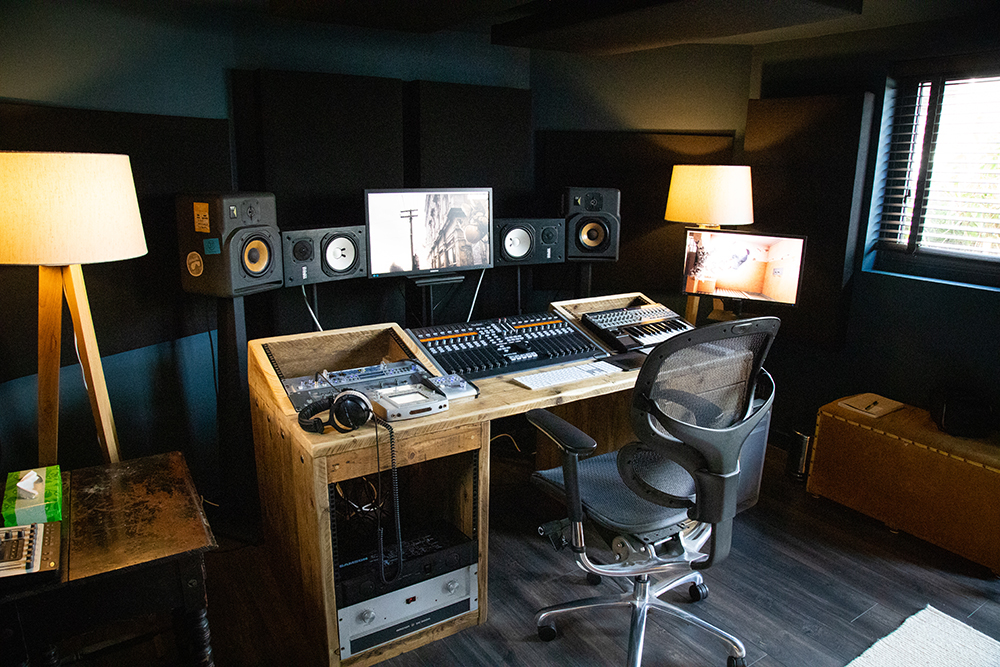 Interior view of a Vivid Green garden music studio's desk featuring technical equipment, screens and speakers and a black office chair, two warm light lamps on either side of the desk, a window to the right of the desk, and a side table on the left.