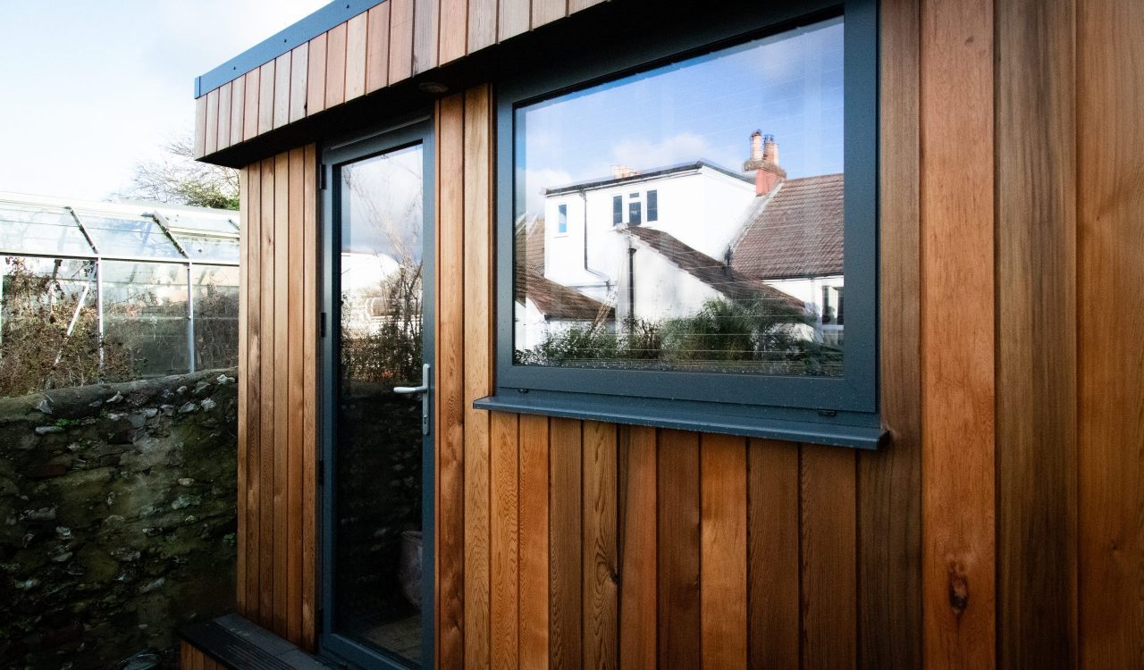 Exterior front view of a Vivid Green, western red cedar clad, music studio space in a back garden featuring an athracite grey door and window with the main house reflecting in the glass