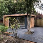 Western red cedar clad Vivid Green garden office space featuring anthracite grey framed windows surrounded by lush foliage with a wooden walkway leading up to the office door.