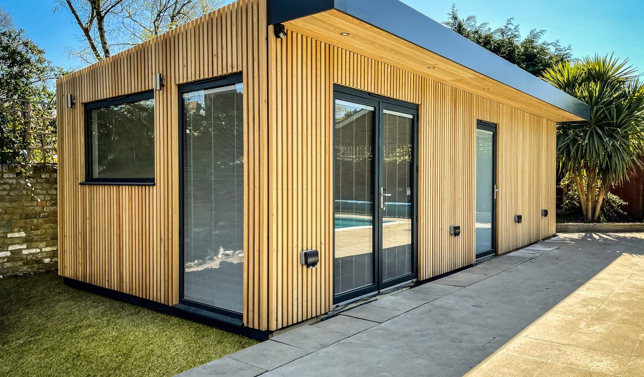 Outside Vivid Green pool room and garden office, larch rain screen cladding, aluminium overhang, anthracite grey framed doors and windows fitted with integral blinds, as well as exterior lights, a tiled patio, and grass located off to the side by a pool