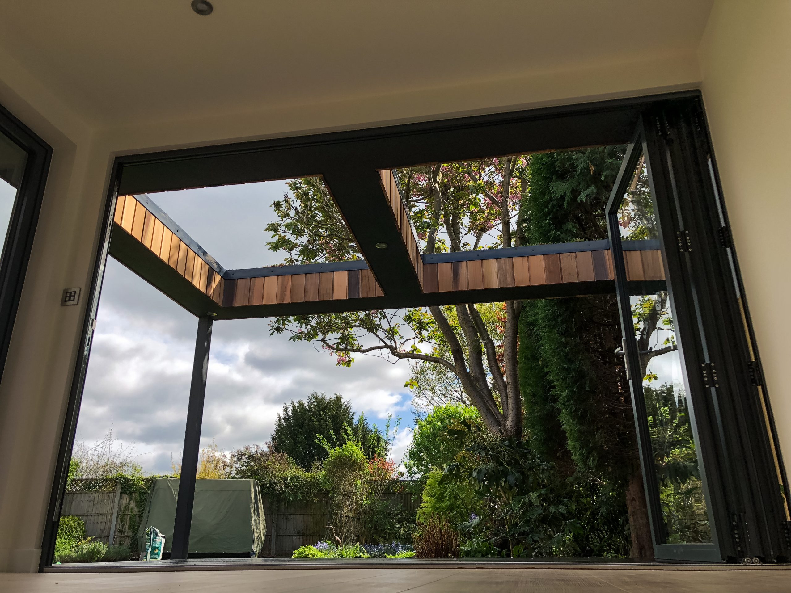 Low angle view from inside a Vivid Green garden office space, featuring anthracite grey aluminum bi-fold doors opening to a cedar and grey trimmed overhang, with the garden in the background.