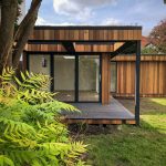 Vivid Green L-shaped garden room with western red cedar cladding, grey composite decking and anthracite grey bi-fold doors and windows, opening to an elevated patio covered by an open overhang, with foliage in the foreground.