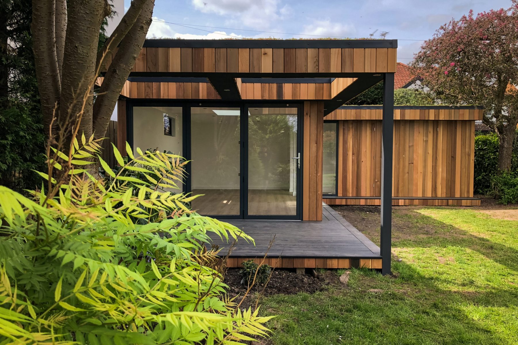 Vivid Green L-shaped garden room with western red cedar cladding, grey composite decking and anthracite grey bi-fold doors and windows, opening to an elevated patio covered by an open overhang, with foliage in the foreground.