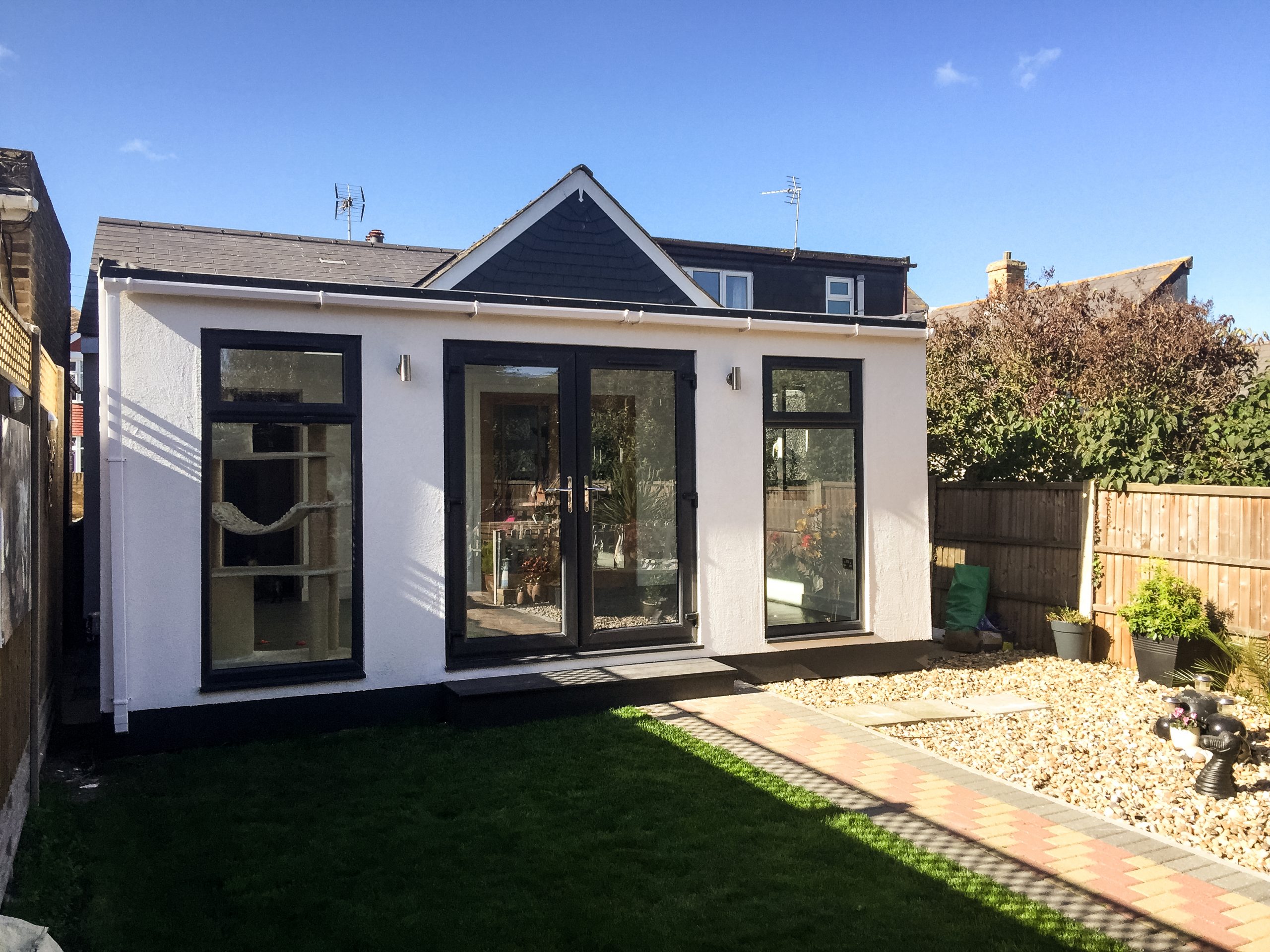 White rendered Vivid Green SIPs house extension with black pitched roof, anthracite grey framed glass door and windows and bricked walkway leading to entrance bordered by gravel and grass