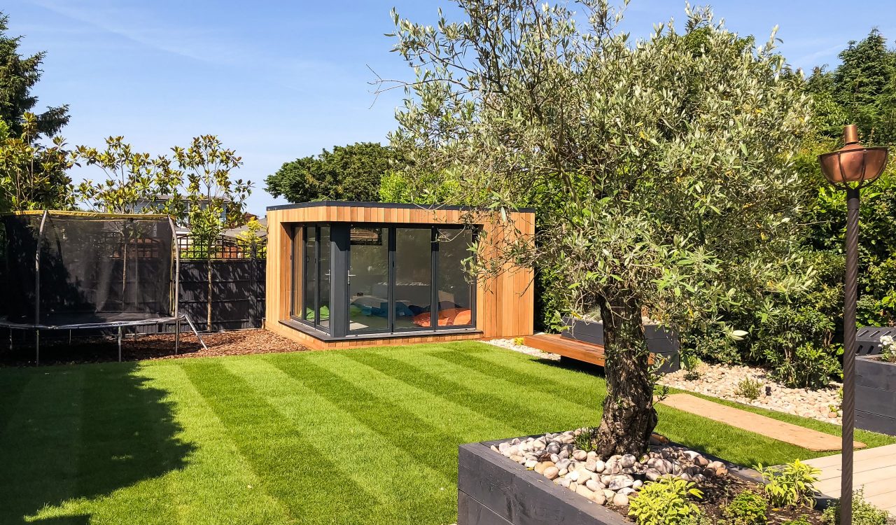 Exterior day view of a small Vivid Green garden office with anthracite grey aluminium framed bi-fold doors with a trampoline to the left of the office and grass garden and steps in the foreground.
